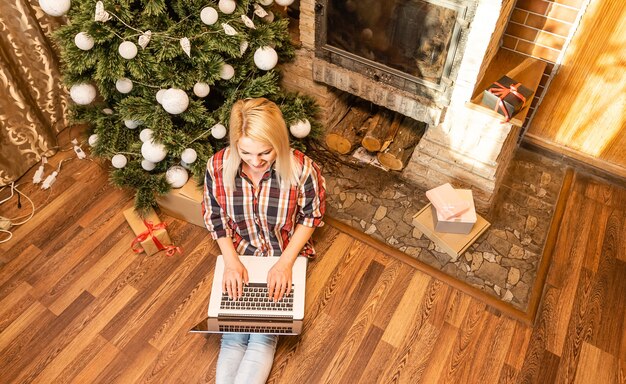 Mujer joven relajante en casa durante la Navidad sentada en el suelo de la sala de estar frente al árbol de Navidad navegando por internet en su computadora portátil.