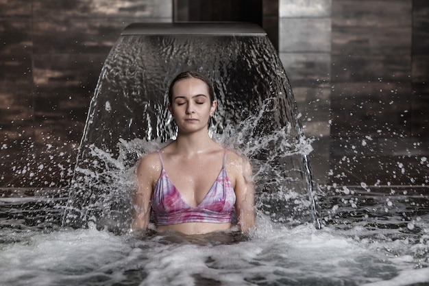 Mujer joven relajante bajo el chorro de agua en la piscina