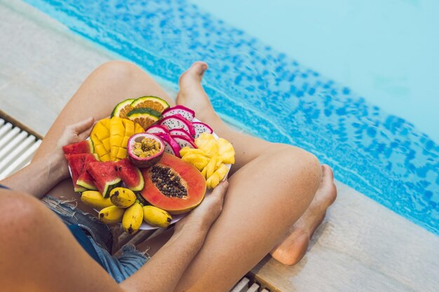 Mujer joven relajarse y comer plato de frutas junto a la piscina del hotel. Dieta exótica de verano. Foto de piernas con comida sana junto a la piscina, vista superior desde arriba. Estilo de vida de playa tropical.