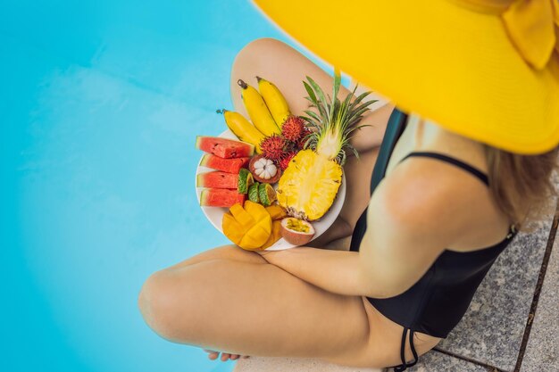 Mujer joven relajarse y comer plato de frutas junto a la piscina del hotel dieta de verano exótica foto de piernas con