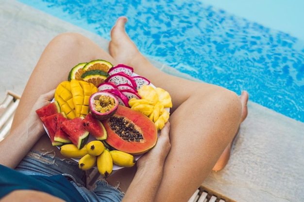 Mujer joven relajarse y comer plato de frutas junto a la piscina del hotel dieta de verano exótica foto de piernas con