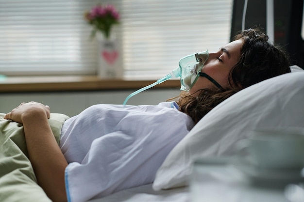 Mujer joven respirando con máscara de oxígeno acostada en la cama durante el tratamiento en el hospital