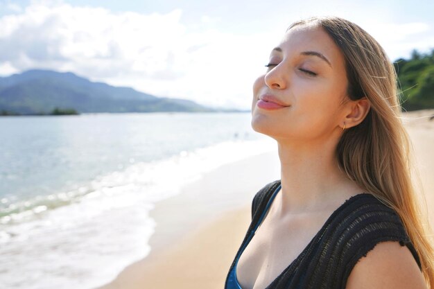 Mujer joven respirando con los ojos cerrados disfruta de tomar el sol en la playa Relax y vacaciones para tomar el sol en la isla de Ilhabela Brasil