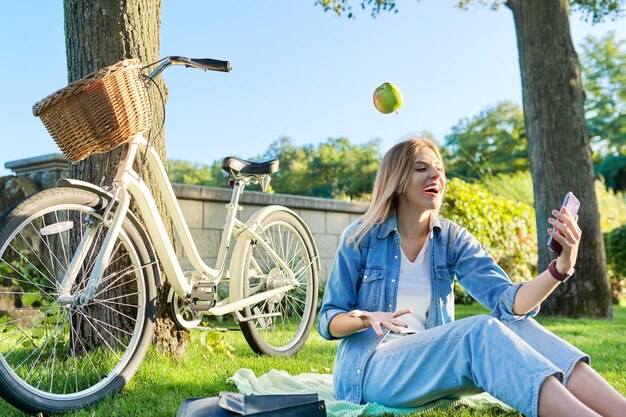 Mujer joven riendo feliz sentada en el césped lanzando manzana haciendo malabares haciendo videollamadas en el teléfono inteligente Mujer hermosa sana activa con bicicleta en el fondo del parque