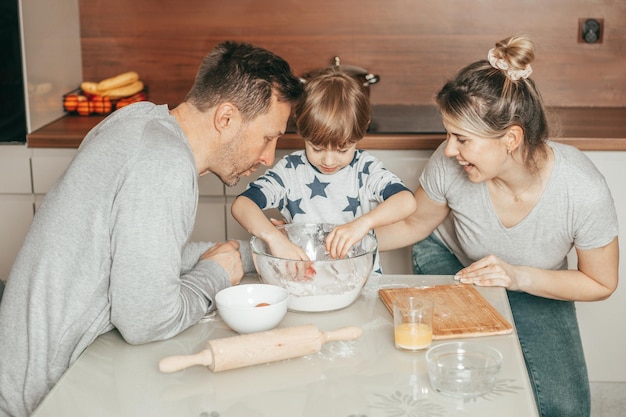 Mujer joven riendo mientras sus hombres, padre e hijo, cocinan galletas juntos. Prepara la masa para hornear. Horas felices de la familia joven. Colaboración como equipo. Los ayudantes de mamá. Cocina casera