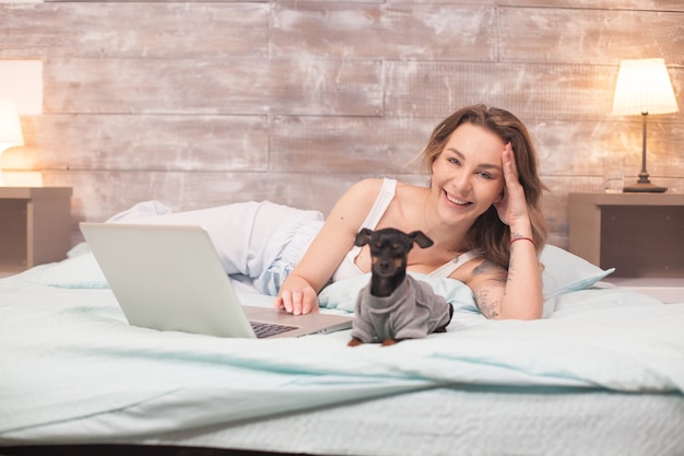 Mujer joven riendo mientras usa su computadora portátil en la cama en pijama. Perrito.