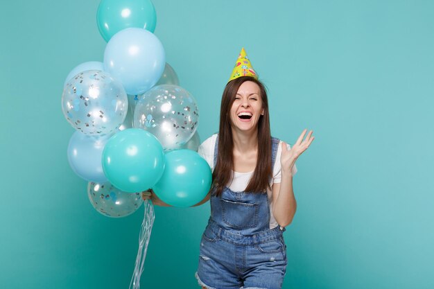 Mujer joven riéndose con ropa de mezclilla, sombrero de cumpleaños esparciendo las manos, celebrando, sosteniendo coloridos globos de aire aislados en un fondo azul turquesa. Fiesta de cumpleaños, concepto de emociones de la gente.