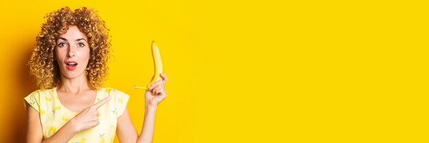 Mujer joven rizada sorprendida sosteniendo un plátano apuntando con su dedo sobre un fondo amarillo Banner