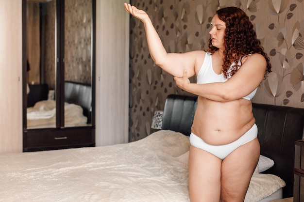 Mujer joven rizada de talla grande en ropa interior blanca parada cerca de la cama en el dormitorio y pellizcando los brazos caídos con la mano Las niñas con sobrepeso luchan contra la obesidad Programa de pérdida de peso control corporal y dieta