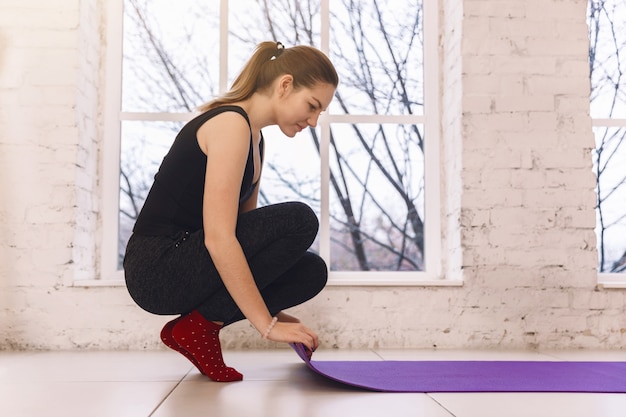 Mujer joven rodando su tapete púrpura después de una clase de yoga en el piso cerca de una ventana,