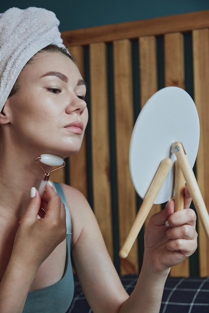 Mujer joven con rodillo facial de jade para masaje facial y de cuello sentada en la cama en el dormitorio mirándose en el espejo