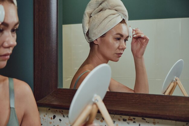 Mujer joven con rodillo facial de jade para masaje facial sentada en el baño mirándose en el espejo