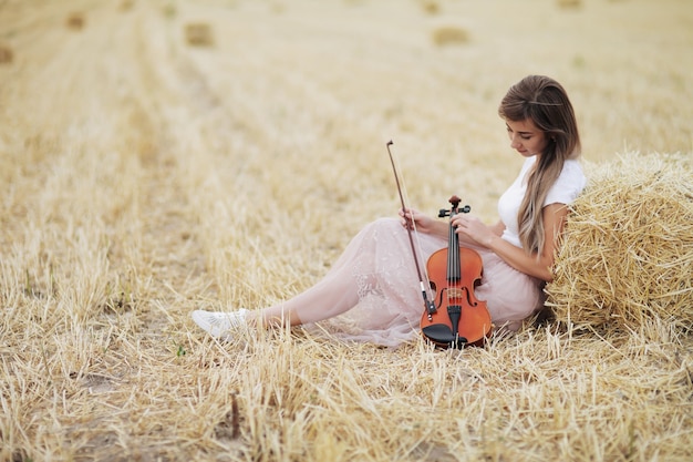 Mujer joven romántica con cabello suelto, sosteniendo un violín en su mano en un campo después de la cosecha