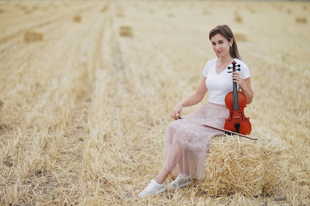 Mujer joven romántica con cabello suelto, sosteniendo un violín en su mano en un campo después de la cosecha