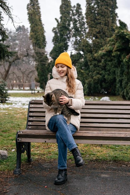 Una mujer joven con ropa de abrigo sostiene un gato y se sienta en un banco del parque Paseando gatos domésticos o ayudando a las personas sin hogar Imagen vertical