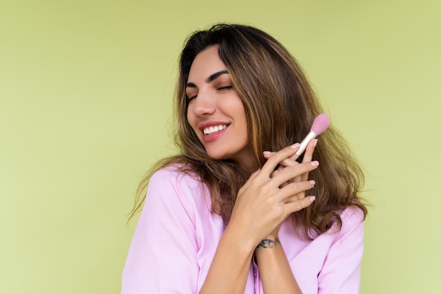 Mujer joven en ropa casual aislada sobre fondo verde juguetona positiva con pincel de rubor maquillaje diario natural