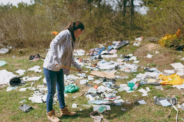 Mujer joven en ropa casual y guantes de látex para limpiar con rastrillo para la recolección de basura en el parque lleno de basura