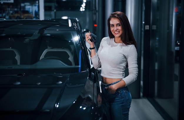 Mujer joven en ropa casual de pie junto a un coche negro moderno con llaves en las manos.