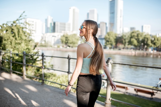 Mujer joven en ropa deportiva caminando y mirando hacia atrás en el parque con rascacielos en el fondo en la ciudad de Frankfurt