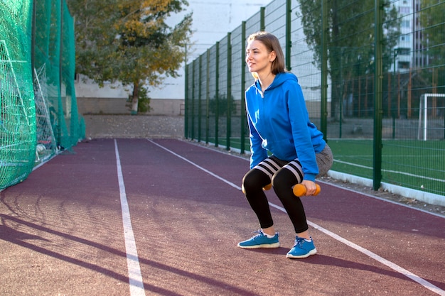 Mujer joven en ropa deportiva está entrenando con pesas en la calle, un estudiante se dedica a deportes al aire libre