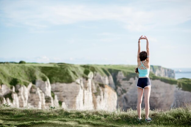 Mujer joven en ropa deportiva estirando al aire libre en el fondo de la hermosa costa rocosa cerca de la ciudad de Etretat en Francia