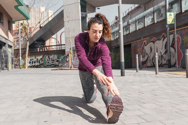 Mujer joven con ropa deportiva estirando la pierna en el suelo al aire libre