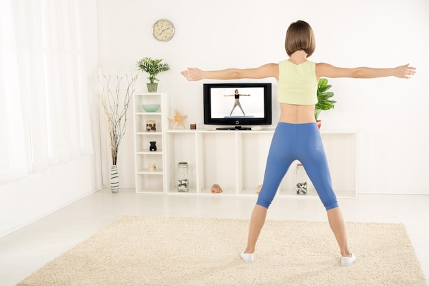 Mujer joven en ropa deportiva, fotografiada desde atrás, ejercicio en la habitación, viendo los ejercicios en la televisión.