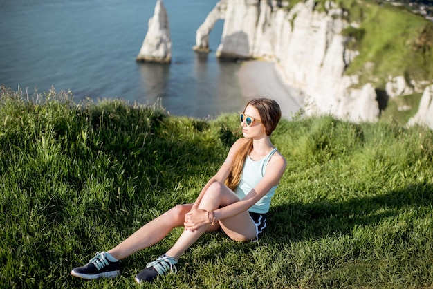 Mujer joven en ropa deportiva sentado al aire libre en el fondo de la hermosa costa rocosa cerca de la ciudad de Etretat en Francia