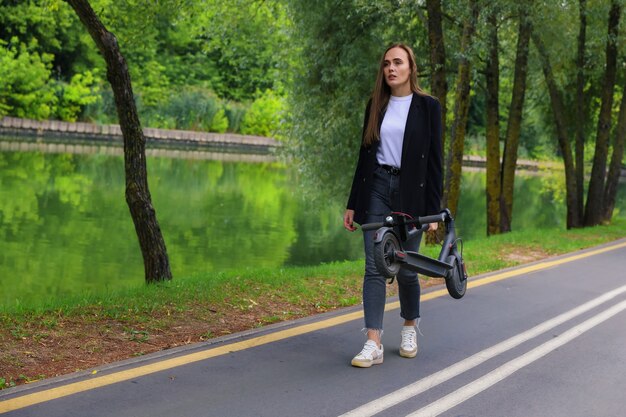 Una mujer joven con ropa elegante camina por un carril bici con un scooter eléctrico plegado