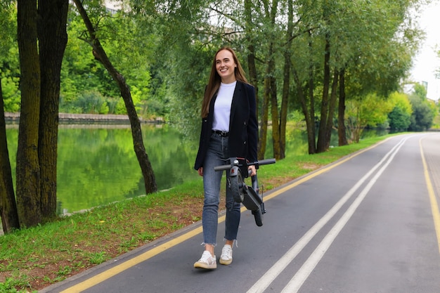 Una mujer joven con ropa elegante camina por un carril bici con un scooter eléctrico plegado