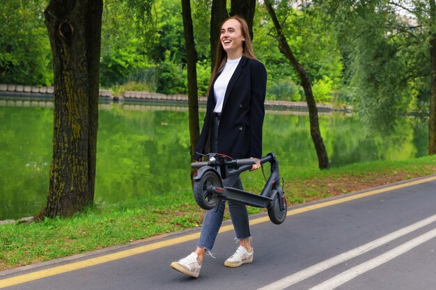 Una mujer joven con ropa elegante camina por un carril bici con un scooter eléctrico plegado