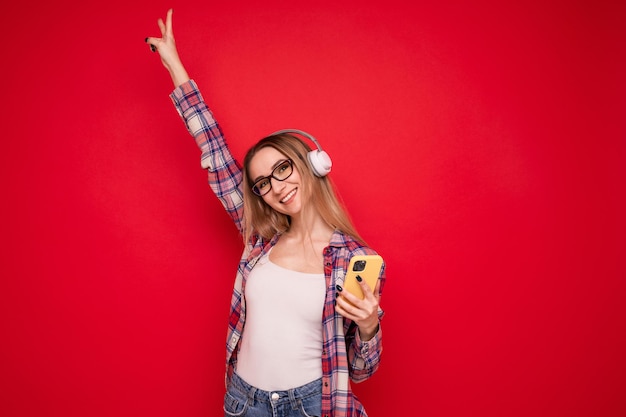 Mujer joven con ropa elegante escucha música con auriculares desde su teléfono en un fondo rojo