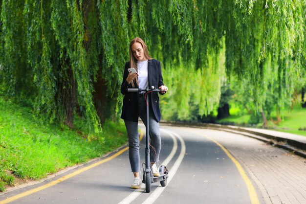 Una mujer joven con ropa elegante posa junto a un scooter eléctrico en un carril bici