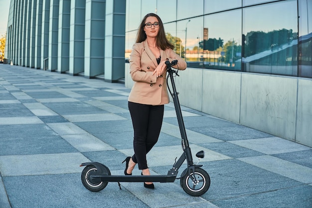 Mujer joven en ropa formal y anteojos está montando scooter eléctrico de su trabajo.