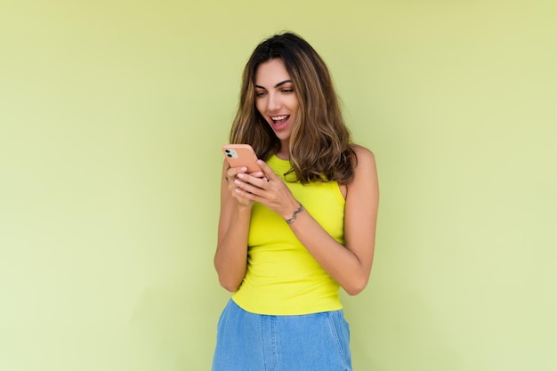 Mujer joven con ropa informal aislada de fondo verde mirando la pantalla del teléfono con una gran sonrisa leyendo el mensaje