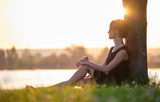 Mujer joven con ropa informal sentada en el césped verde relajándose al lado del lago en una noche cálida Vacaciones de verano y concepto de viaje