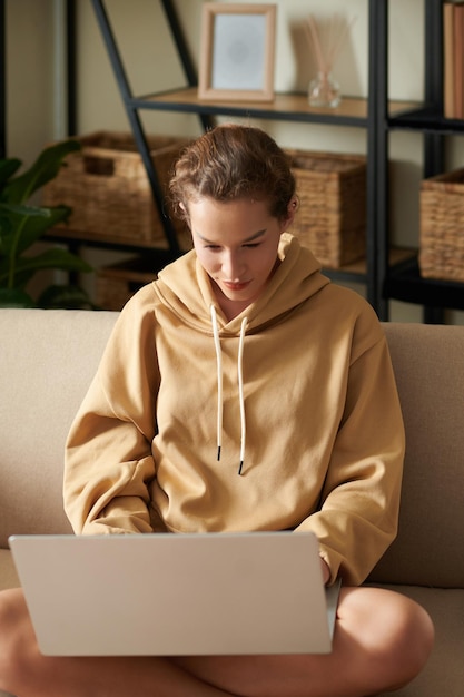 Mujer joven con ropa informal sentada en un sofá en la habitación con una laptop de rodillas y concentrándose en su trabajo en línea