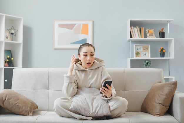 mujer joven con ropa informal sentada en un sofá en el interior de su casa usando un teléfono inteligente mirando la pantalla sorprendida y confundida levantando el brazo