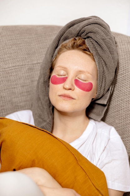 Mujer joven con ropa informal toalla en la cabeza y parches en los ojos sentada en el sofá y descansando Relájese después de ducharse Cosmética para el cuidado de la piel de las mujeres Tiempo de autocuidado para usted Relajarse en casa
