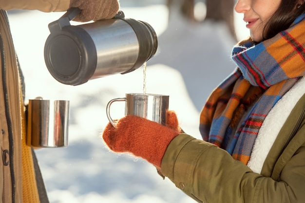 Mujer joven en ropa de invierno abrigada sosteniendo una taza metálica mientras su marido sirve té caliente del termo frente a la cámara contra la nieve