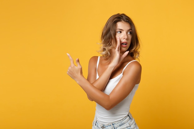 Mujer joven en ropa ligera posando aislada sobre fondo amarillo naranja. Concepto de estilo de vida de las personas. Simulacros de espacio de copia. Susurra chismes y cuenta secretos con un gesto de la mano, señalando con el dedo índice a un lado.