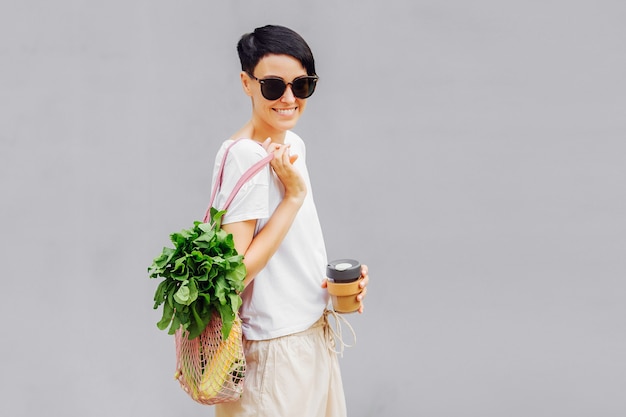 Mujer joven en ropa ligera de verano con una bolsa ecológica de verduras, verduras y taza de café reutilizable. Estilo de vida sostenible. Concepto ecológico.