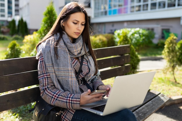 Una mujer joven con ropa de otoño está trabajando atentamente en una computadora portátil mientras está sentada en un banco afuera