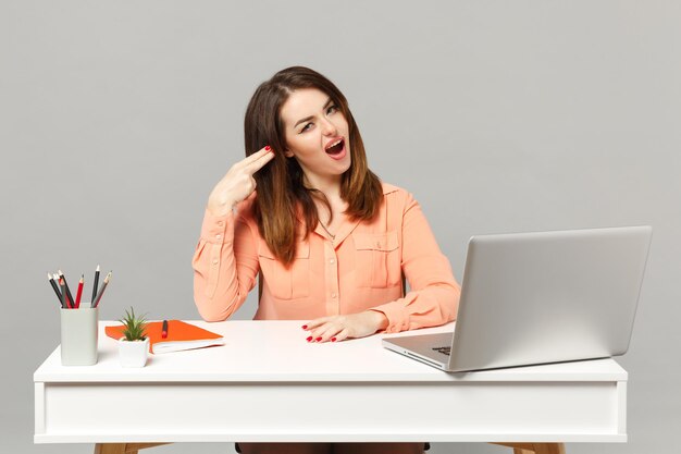 Mujer joven con ropa pastel señala con el dedo a la cabeza como si se disparara a sí misma sentada en el escritorio con una computadora portátil aislada en un fondo gris. Logro el concepto de carrera empresarial. Simulacros de espacio de copia.