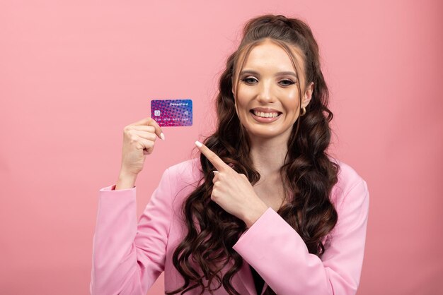 Mujer joven con ropa rosa sonriendo sosteniendo una tarjeta de crédito de plástico cerca de la cara y mirando