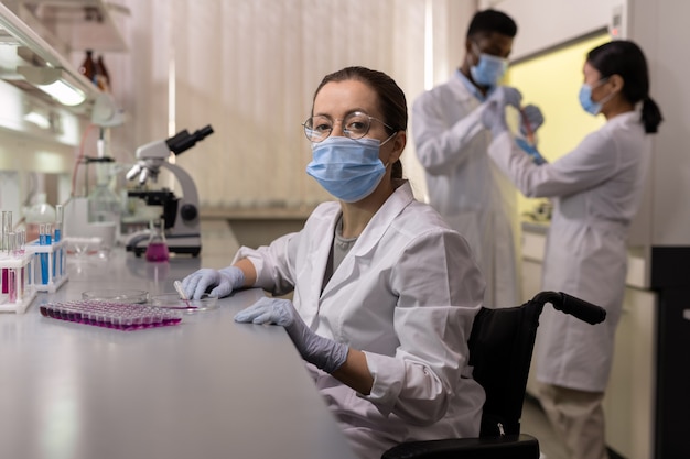 Mujer joven en ropa de trabajo protectora sentada por lugar de trabajo en laboratorio médico