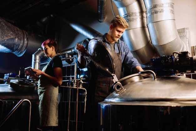 Mujer joven en ropa de trabajo con tableta digital mientras su colega masculino abre la tapa de la enorme cisterna de acero durante el proceso de producción de cerveza