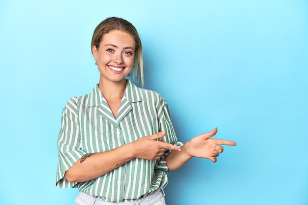 Foto mujer joven rubia con camisa a rayas verdes sobre fondo azul emocionada señalando con los dedos índice lejos