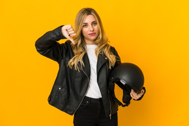 Mujer joven rubia caucásica del motorista que sostiene el casco que muestra un gesto de disgusto, pulgares hacia abajo. Concepto de desacuerdo.