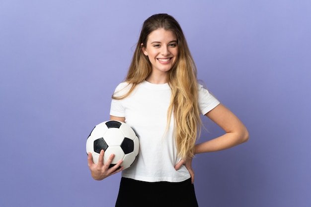 Mujer joven rubia jugador de fútbol aislada en el espacio púrpura posando con los brazos en la cadera y sonriendo